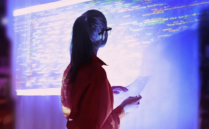 A woman analyzing codes projected on the wall carrying a paper in her hands.