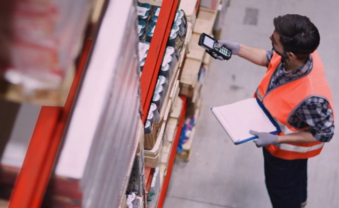 A warehouse worker using a handheld barcode scanner while holding a clipboard in an aisle stocked with palletized goods.