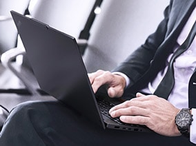 Business professional in a suit working on a laptop while seated in an airport terminal, with a carry-on suitcase beside him.