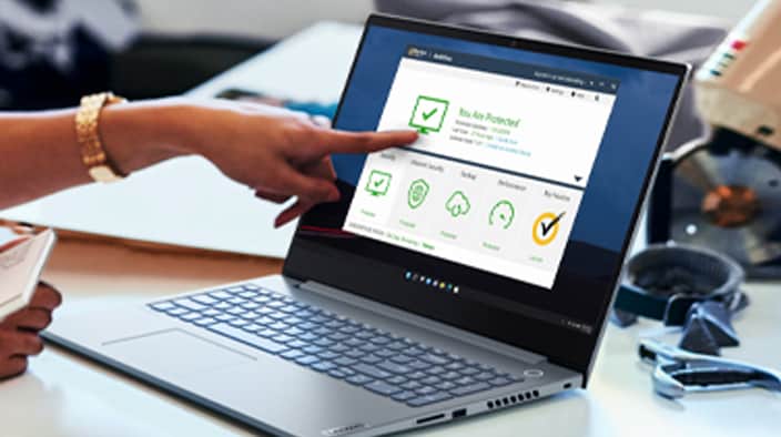The image shows a person pointing at the screen of an open Lenovo laptop. The screen displays a Norton security software interface with checkmark icons. The pointing hand indicates interaction with the software, and scattered objects on the table suggest an active and creative workspace.