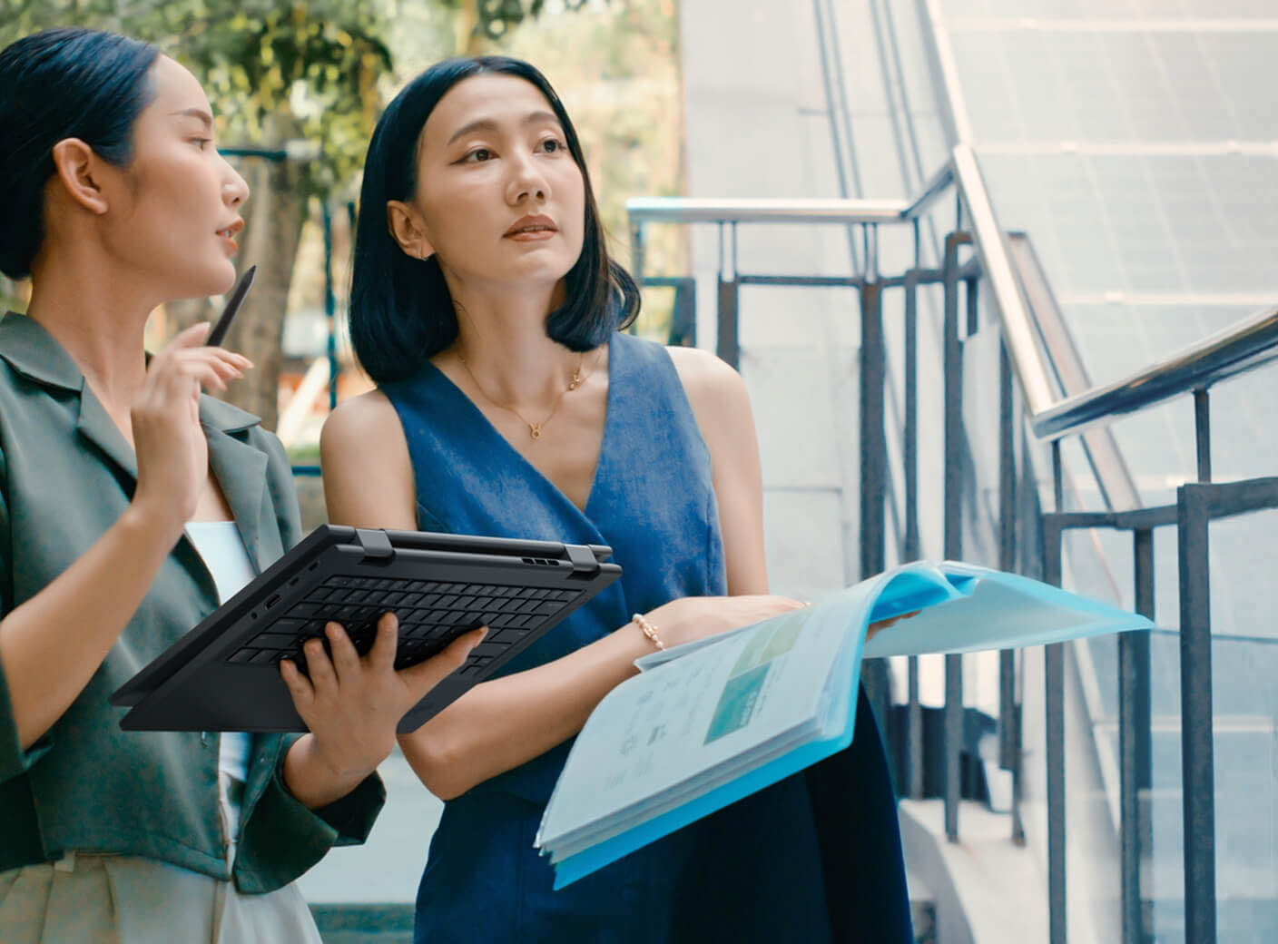 Two women working outdoors, one holding a tablet and the other holding a paper notebook.