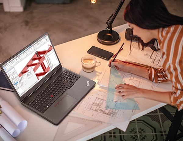 Aerial view of architect at work, with Lenovo ThinkPad P16v (16” AMD) mobile workstation showing designs in screen, and paper drawings/designs on desk