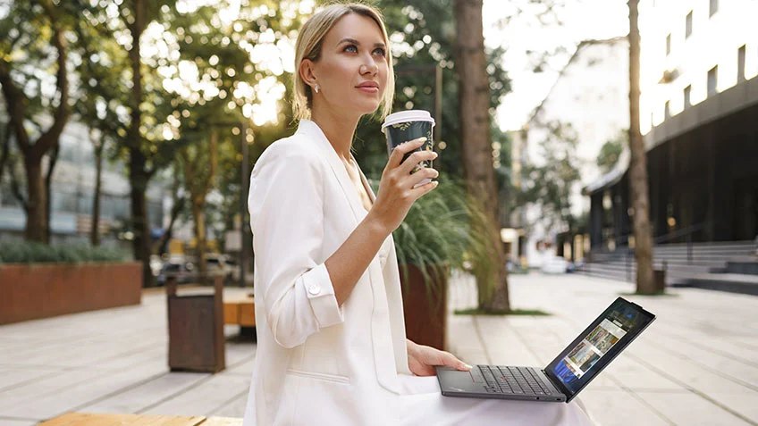 Eine Frau sitzt draußen auf einer Bank. Ihr Notebook ist auf dem Schoß geöffnet und sie hält einen Kaffeebecher in der Hand.