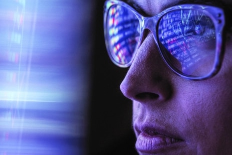 A close-up of a woman's face and the code from a computer screen is reflected in her glasses