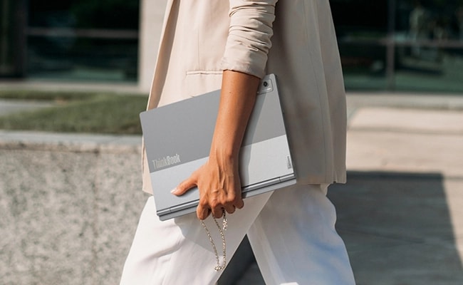 Cut-short view of a person carrying Lenovo ThinkBook Plus Gen 5 Hybrid (14” Intel) laptop, closed, with visible dua-camera setup & ThinkBook logo, boasting its portability.