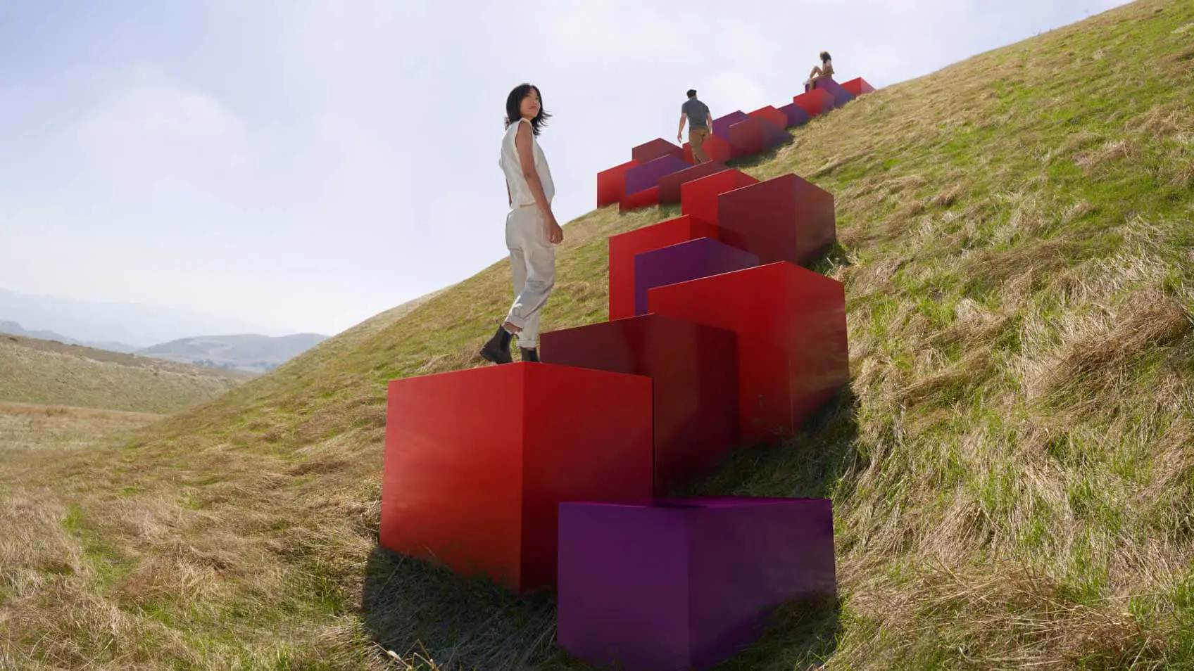 Red and purple cubes leading up a grassy hill with three people ascending them