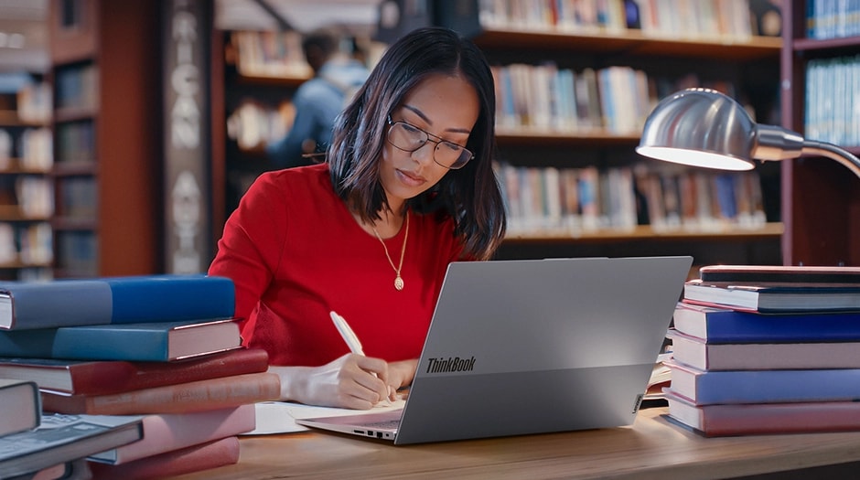 A woman sits at a desk with books in a library writing notes & using a Lenovo ThinkBook 16 Gen 9 laptop.