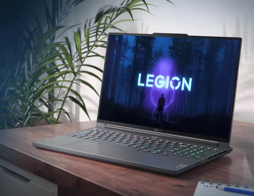 Three-quarters left-facing mid-shot of Lenovo Legion Slim 7 sitting on a desk with the Legion logo visible on the display and plants to the left of the desk in the background.