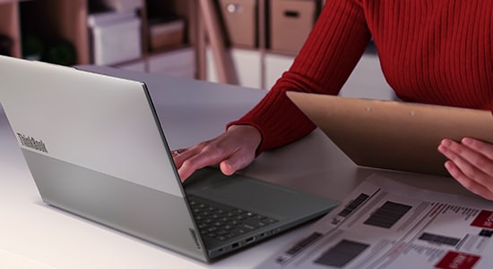 A person sits with a Lenovo ThinkBook laptop on their lap, dressed in a purple skirt and blue top, holding a red textured mug in one hand. The scene is well-lit with natural light, and the laptop lid is partially open, showcasing its sleek, silver-gray design and subtle branding.