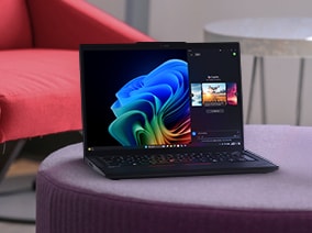 A sleek black laptop displaying a vibrant Windows desktop screen with AI-powered features, placed on a purple ottoman in a modern room with a red chair in the background.