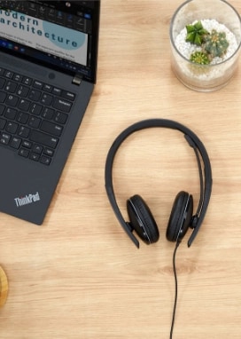 A pair of Lenovo wired headphones on a wooden desk next to a Lenovo ThinkPad X1 Carbon laptop.