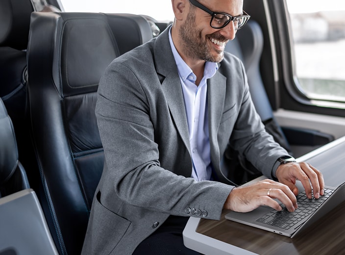 The image shows a person working on a laptop while seated in a modern, well-lit train or similar transport setting.