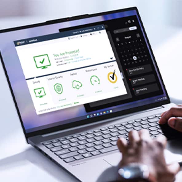 The image shows a person pointing at the screen of an open Lenovo laptop. The screen displays a Norton security software interface with checkmark icons. The pointing hand indicates interaction with the software, and scattered objects on the table suggest an active and creative workspace.