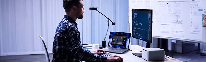 Person seated at a desk working on a laptop and an external monitor displaying code, with a desk lamp and a small desktop device, in a modern office setting with white vertical blinds and technical diagrams on the wall.