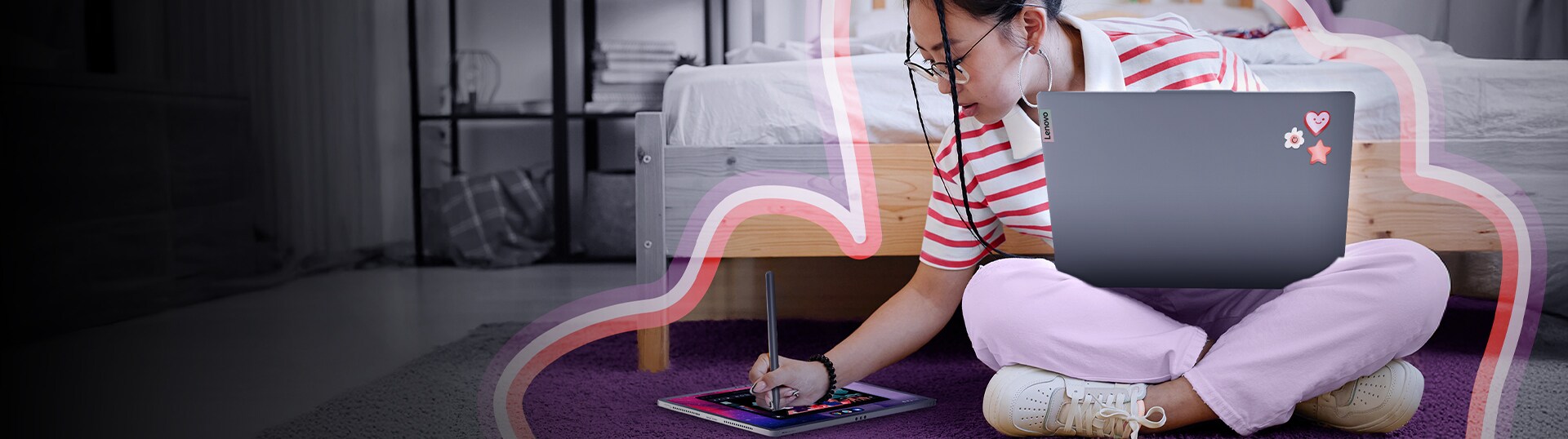 A young woman sitting cross-legged on the floor in a bedroom, working or drawing on a tablet with a stylus. She has a Lenovo laptop resting on her lap, decorated with stickers.