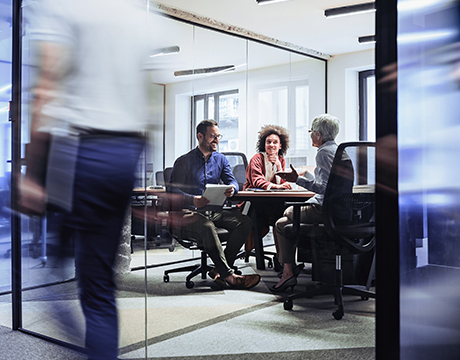 Three people in a glass-walled office meeting space discuss at a table, with blurred motion of a walking person.