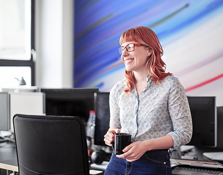 A smiling woman holds a mug standing in an office, highlighting comfort working on the Lenovo ThinkPad P14s Gen 7 (14" AMD) laptop placed behind her.