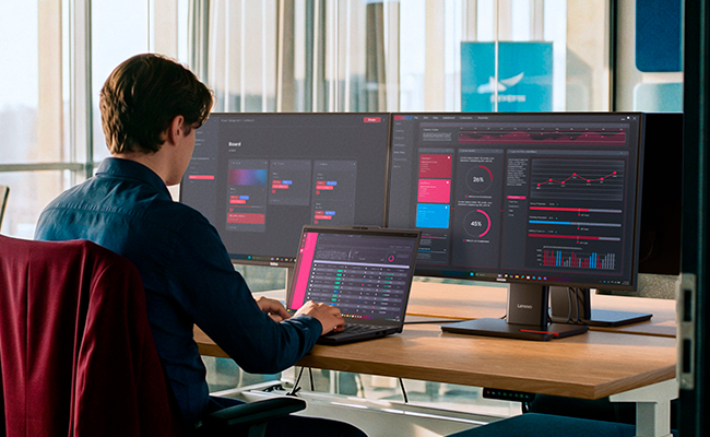 A man at a desk with two monitors types on a Lenovo ThinkPad T14 Gen 7 (14” AMD) laptop.