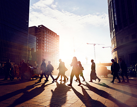 Crowd walking through a sunlit cityscape with tall buildings in the background.
