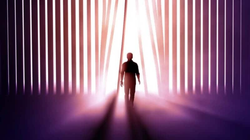 Silhouette of a man walking through an opening in high vertical blinds and into a brightly lit room.