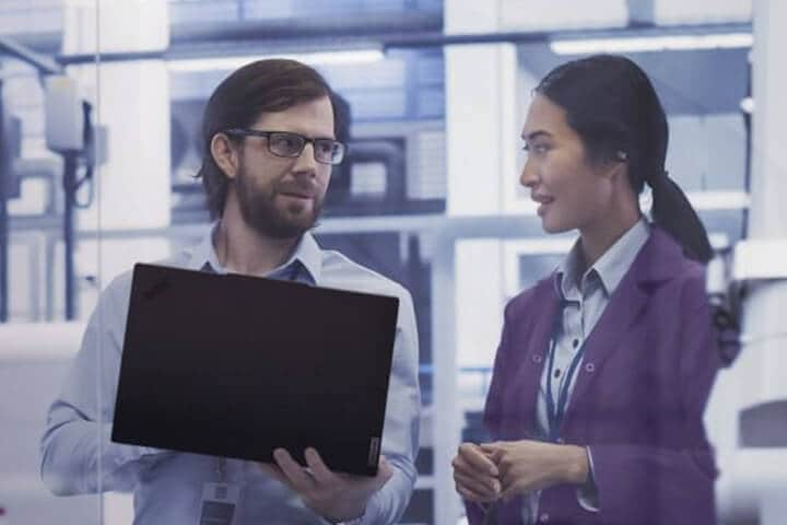 A man holding an open laptop in his hand is speaking with a woman while standing inside an office environment.