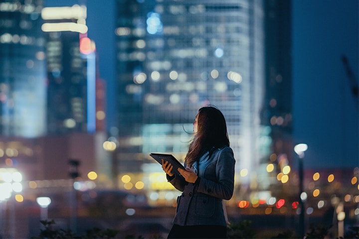 Image shows a woman holding a tablet while looking over her shoulder to a cityscape in the background.