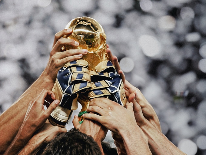 Close-up of multiple football/soccer players' hands holding aloft a FIFA World Cup trophy.