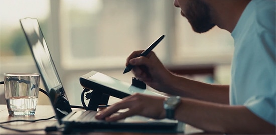 Seated at a desk, Vexx simultaneously types on a Yoga laptop and draws on a tablet. 
