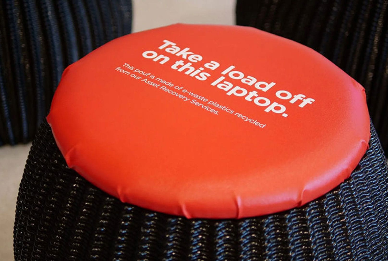 A pouf made of recycled e-waste plastics with a sign on tops that says “Take a load off on this laptop”