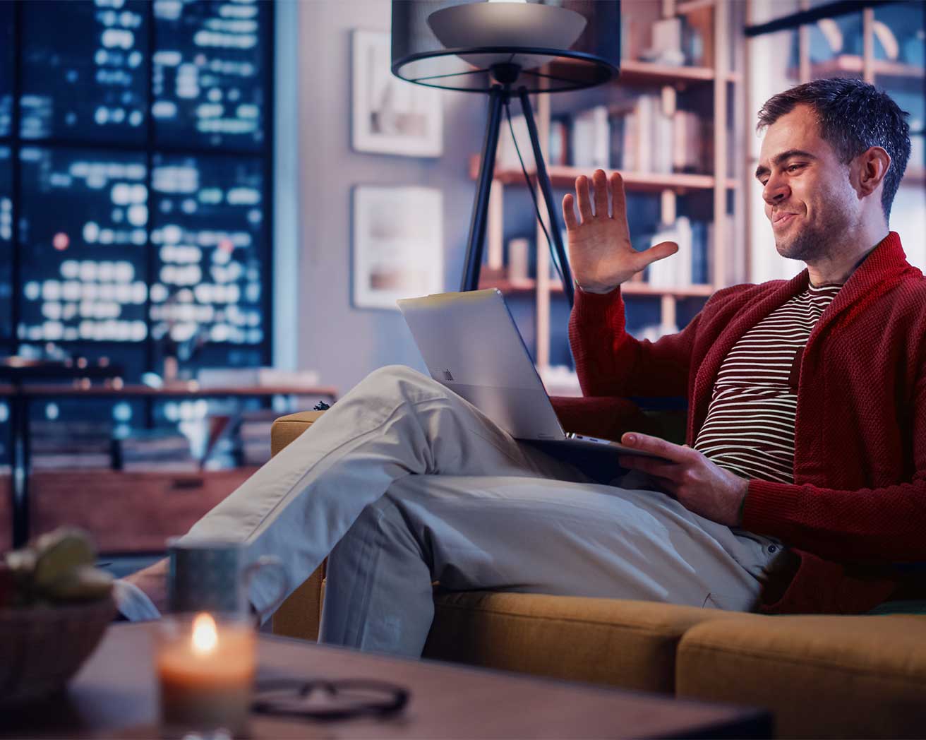 A man sitting on a couch and waving hi to the video chat on his laptop screen.