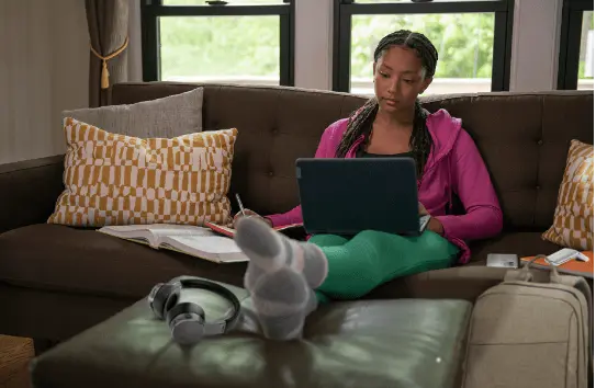 A woman sits on a couch, textbook to her side and a laptop on her lap, feet up on an ottoman, and takes notes while looking at a Lenovo laptop