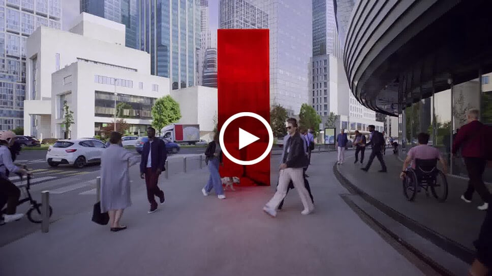 A foto mostra uma rua movimentada com pedestres andando em uma calçada onde há um painel vermelho marcante