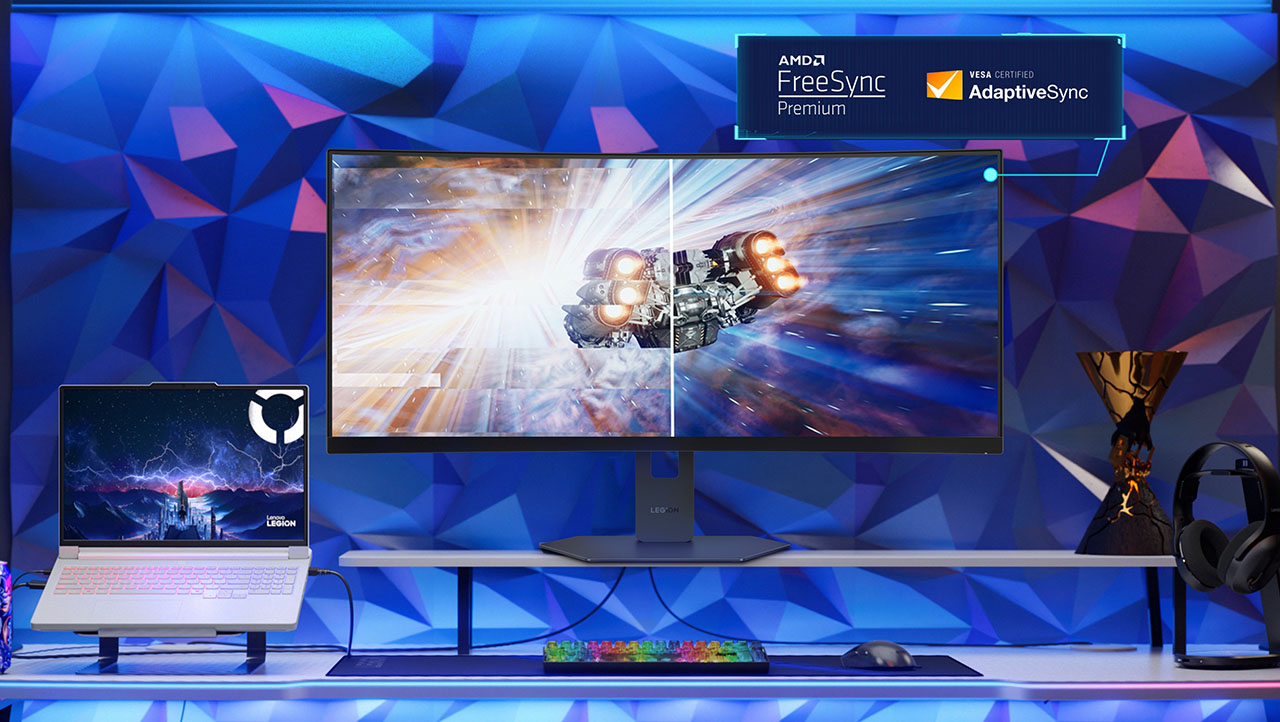 Legion 34w-2c gaming monitor (AMD FreeSync Premium, VESA AdaptiveSync) displaying a split-screen space battle, paired with a Legion laptop, RGB keyboard, mouse, and headphones on a blue geometric desk setup.