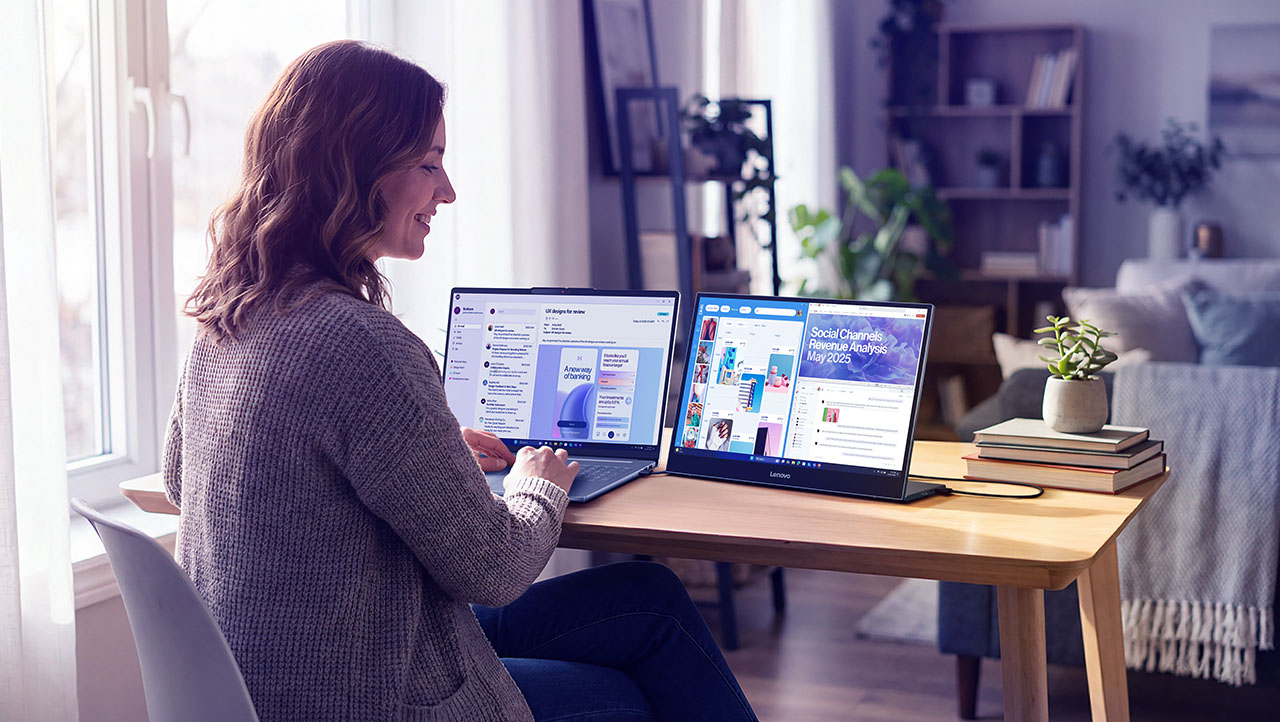 A woman sits at a desk using two laptop screens in a bright, modern home office with books and a potted plant on the table.