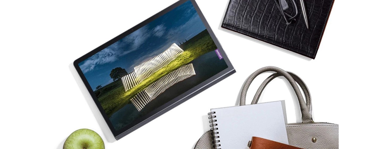 Lenovo Yoga Tab 11 tablet—front view, lying flat on a surface along with an apple, a notebook, a purse, a pair of glasses, and other personal items