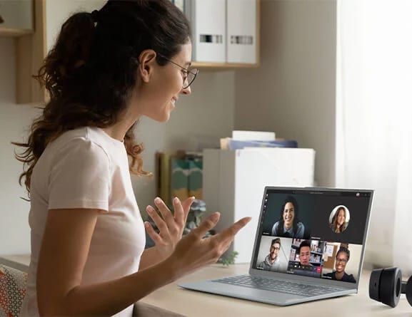 A woman smiles and gestures with her hands as she looks at a Lenovo ThinkBook 13x laptop on a desk in front of her, where five colleagues are shown on-screen in a group video conference.